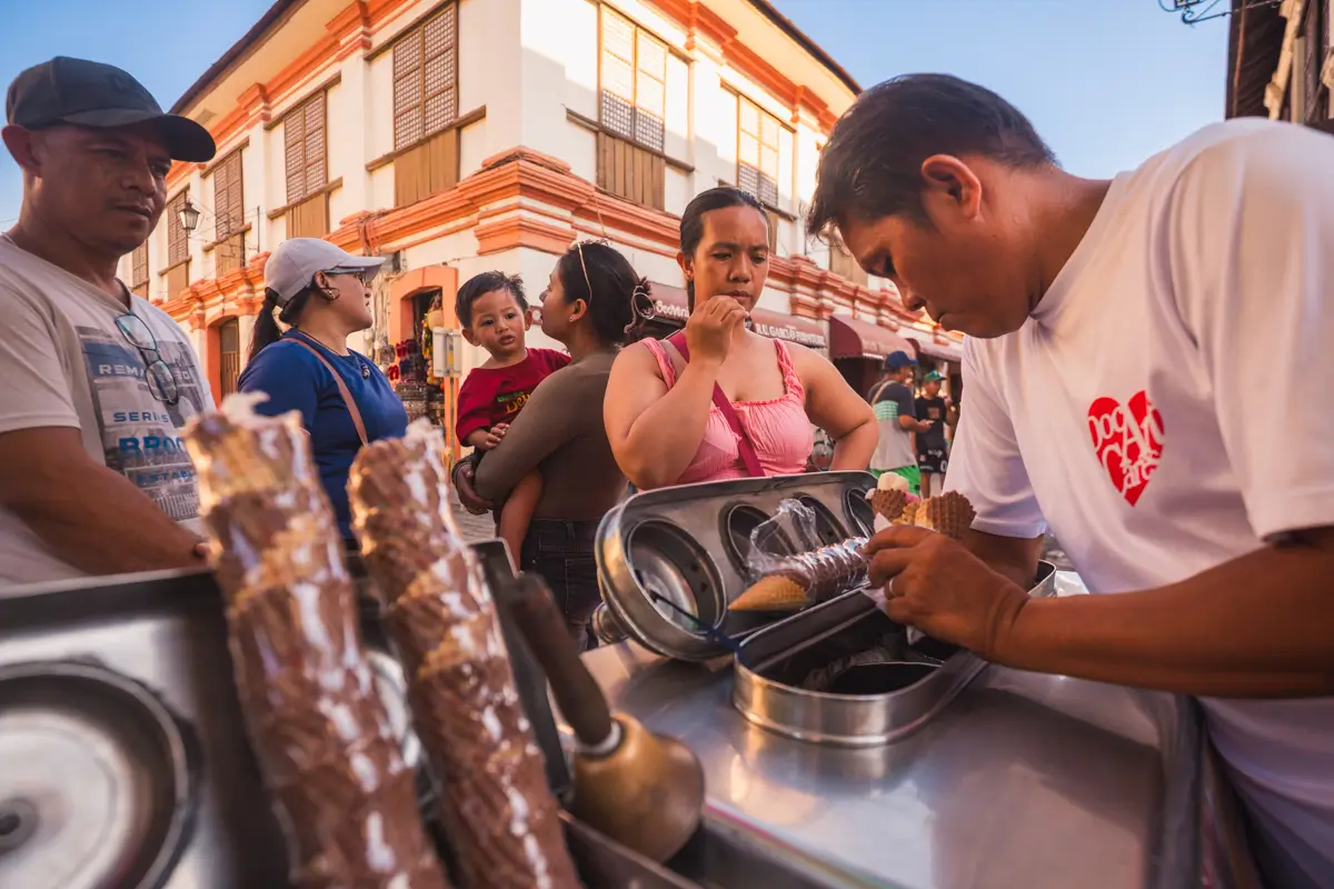 A man in a white shirt prepares ice cream cones at a street cart while several people, including a child, wait in line on a sunlit street with colonial-style buildings—a classic scene from Vigan Tourist Spots.