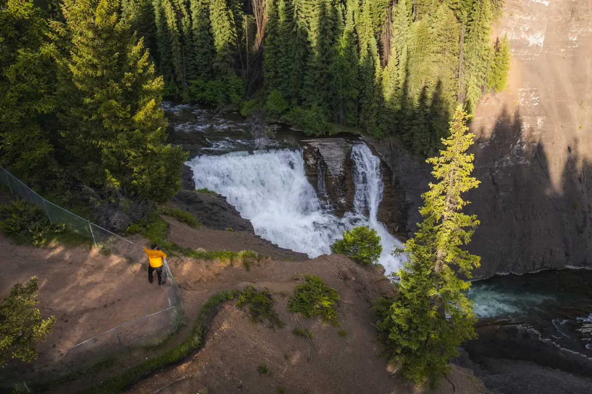 A person in a yellow jacket stands on a dirt path near the edge of a cliff, overlooking a waterfall surrounded by dense evergreen trees under sunlight in Fernie.