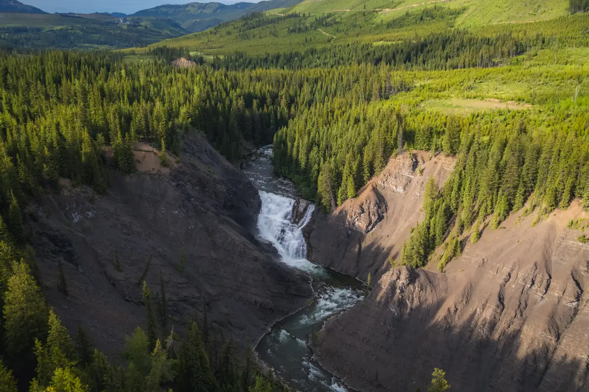 A river flows through a forested Fernie valley with a waterfall cascading between steep, rocky cliffs, surrounded by dense green trees and rolling hills under a clear sky.