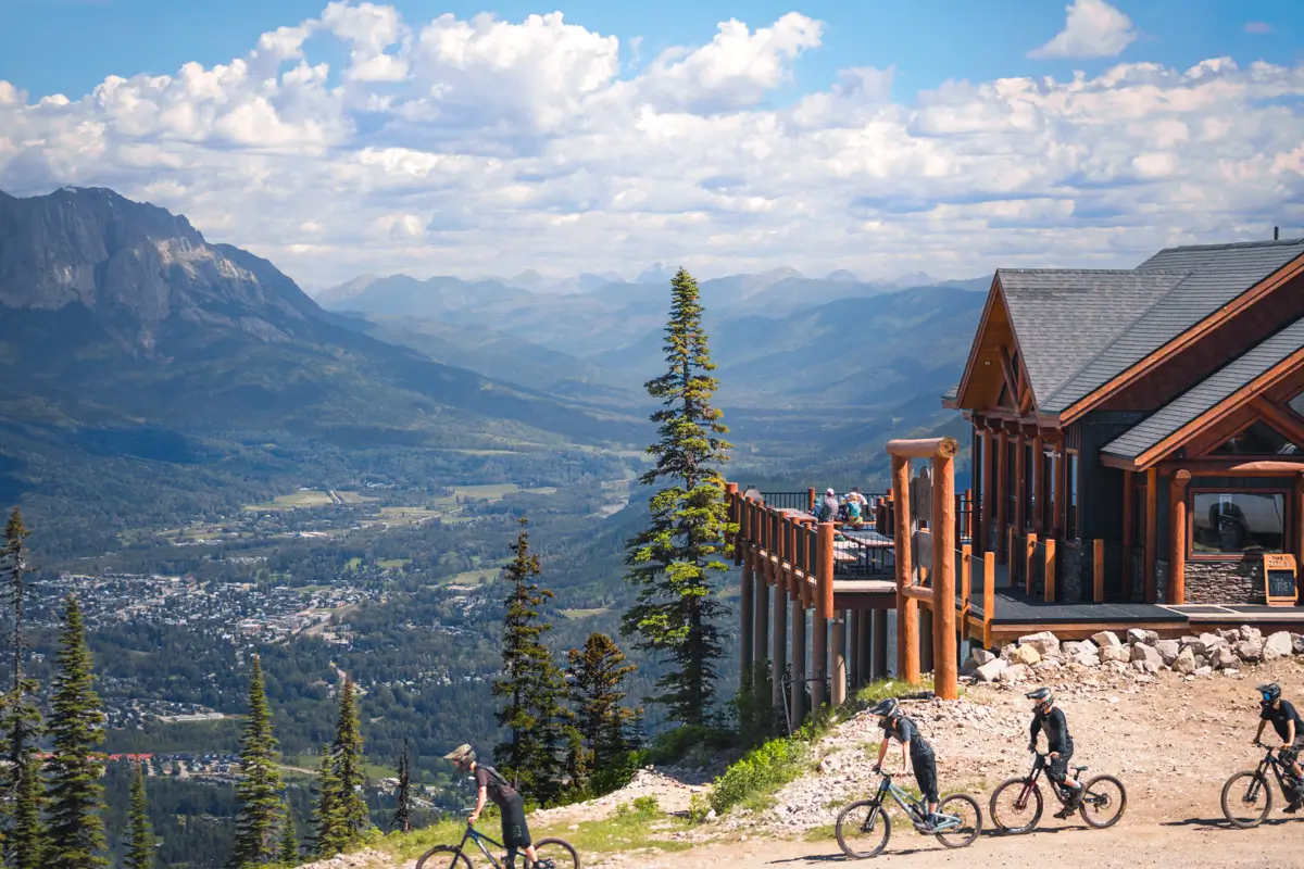 Several cyclists ride on a mountain trail near a wooden lodge with large windows in Fernie, surrounded by pine trees and overlooking a valley, a distant town, and mountain range under a partly cloudy sky.