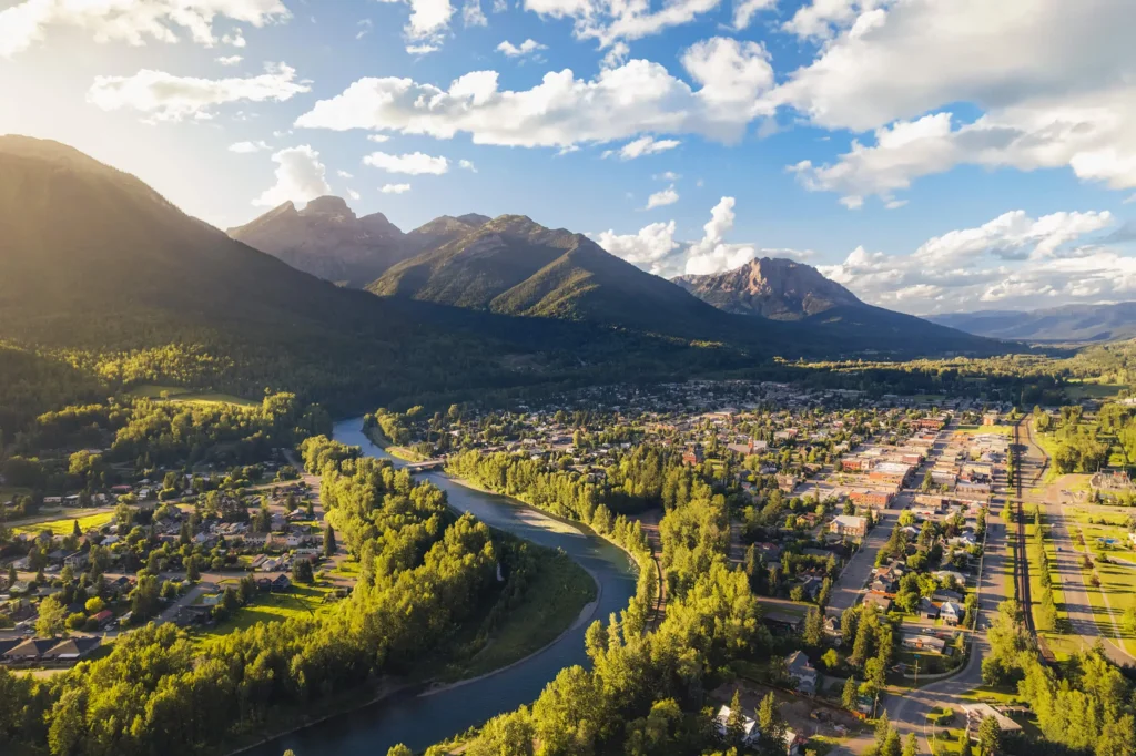 A river curves through the small town of Fernie, with rows of houses surrounded by dense green forests and tall mountains under a partly cloudy sky, sunlight illuminating the landscape.