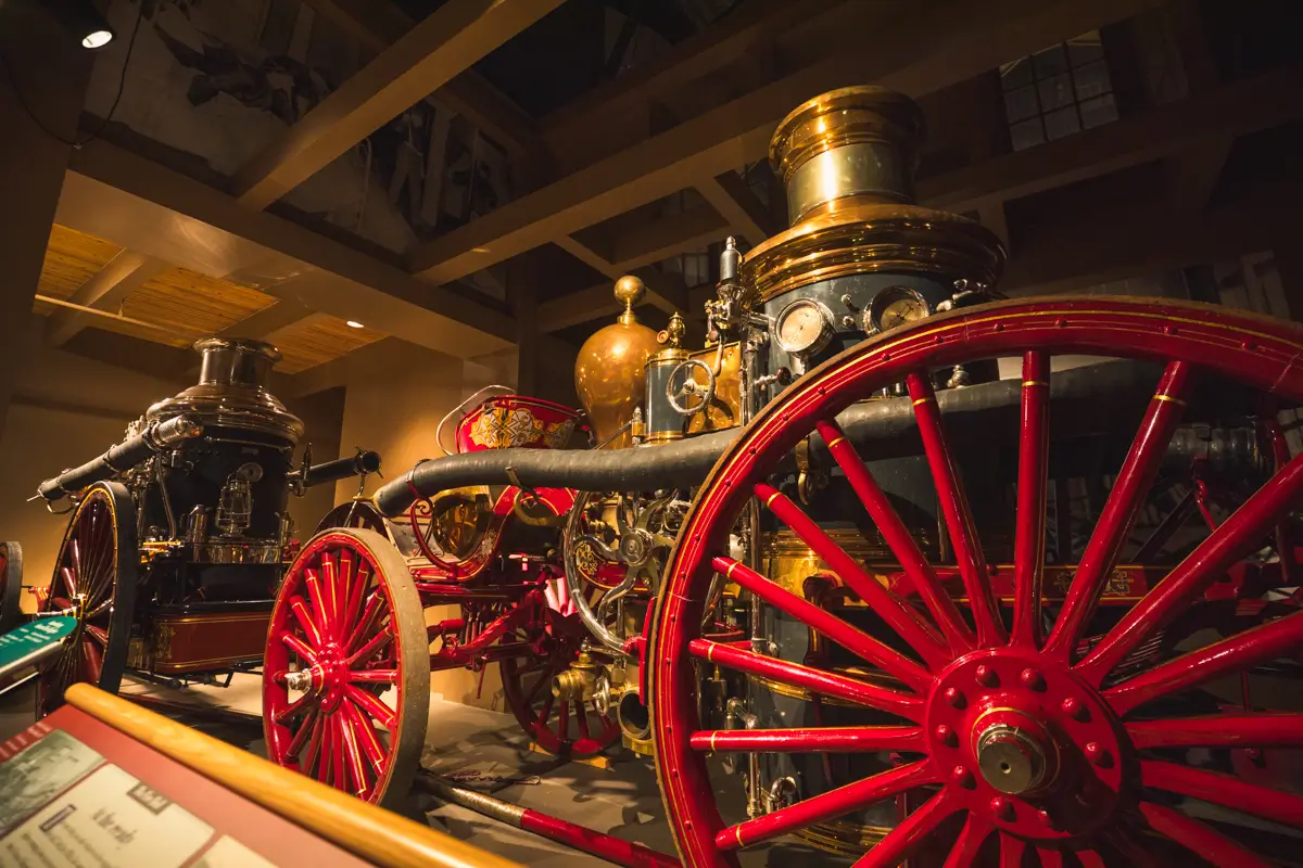 A historic steam-powered fire engine with red wheels and brass details is on display indoors under a wooden ceiling in Lethbridge, alongside another similar antique vehicle.