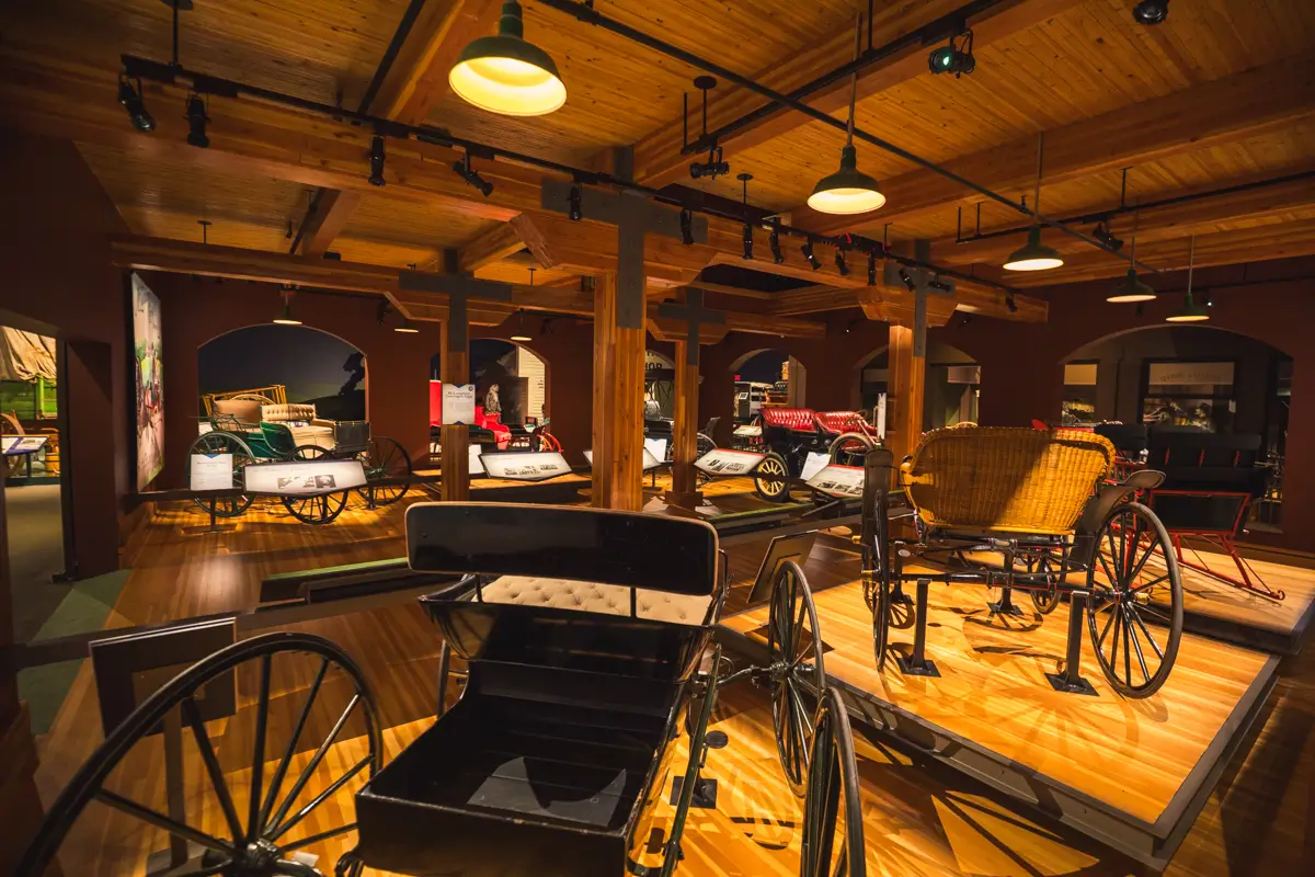 A museum exhibit in Lethbridge displays several antique carriages on raised wooden platforms under warm lighting, with wooden beams and columns supporting the ceiling.