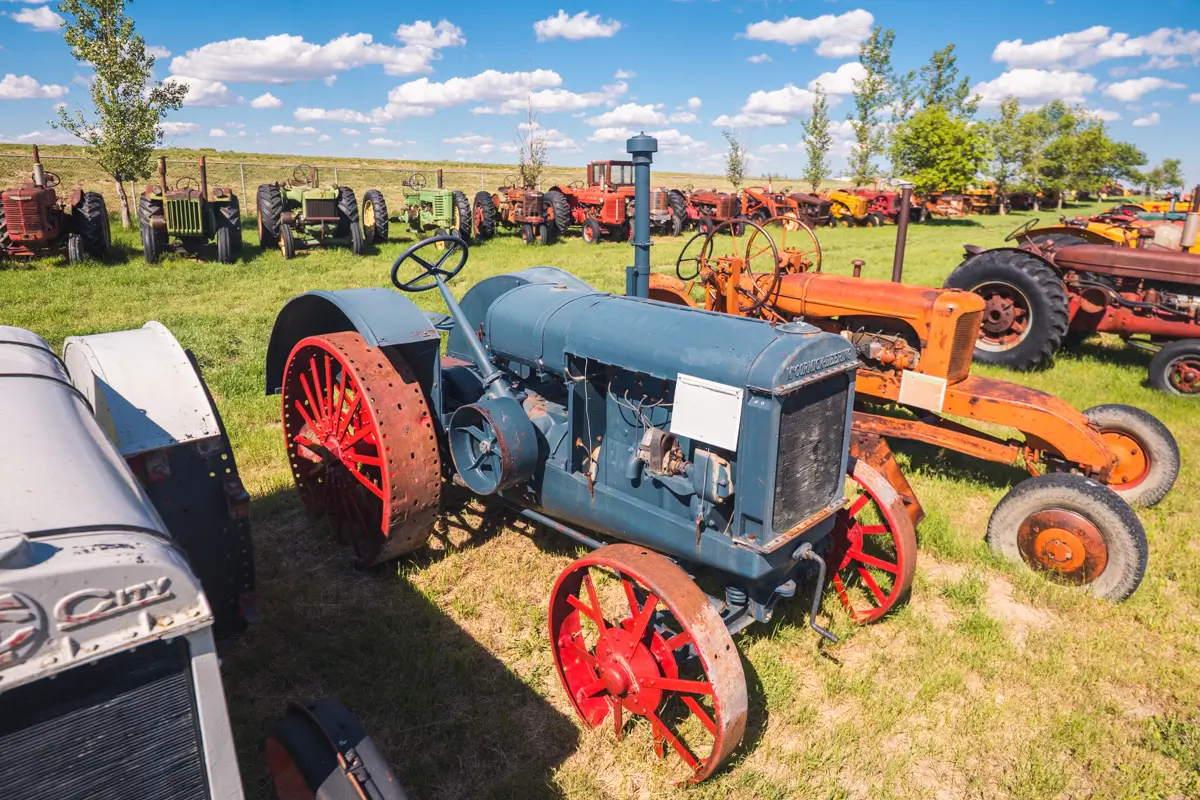 A collection of vintage tractors is displayed outdoors on a grassy field under a blue sky with scattered clouds in Lethbridge, with one blue tractor featuring red wheels prominently in the foreground.