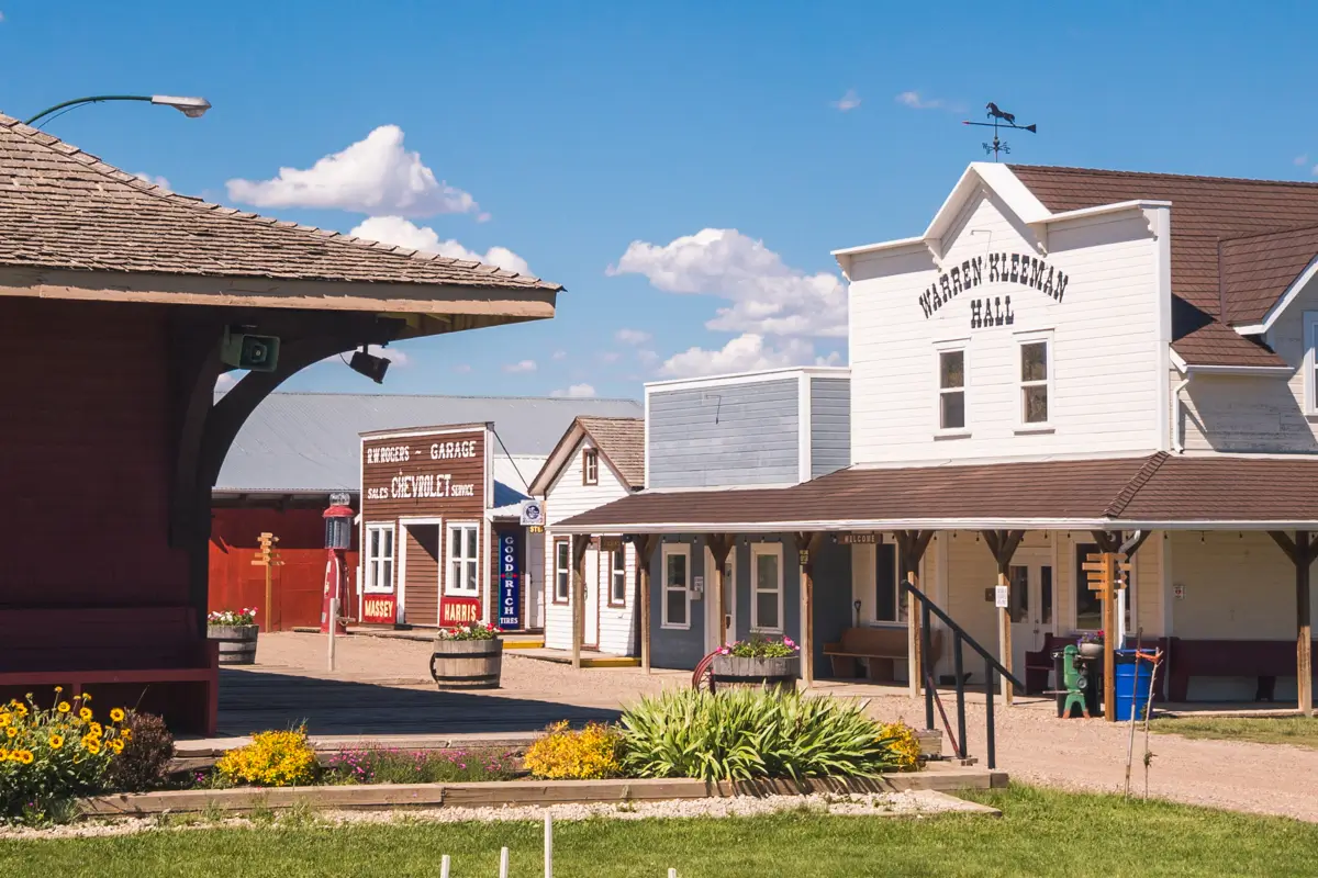 A small Western-style town scene in Lethbridge, featuring wooden buildings—one labeled Hershey American Hall—boardwalks, flower beds, and a bright blue sky with scattered clouds.