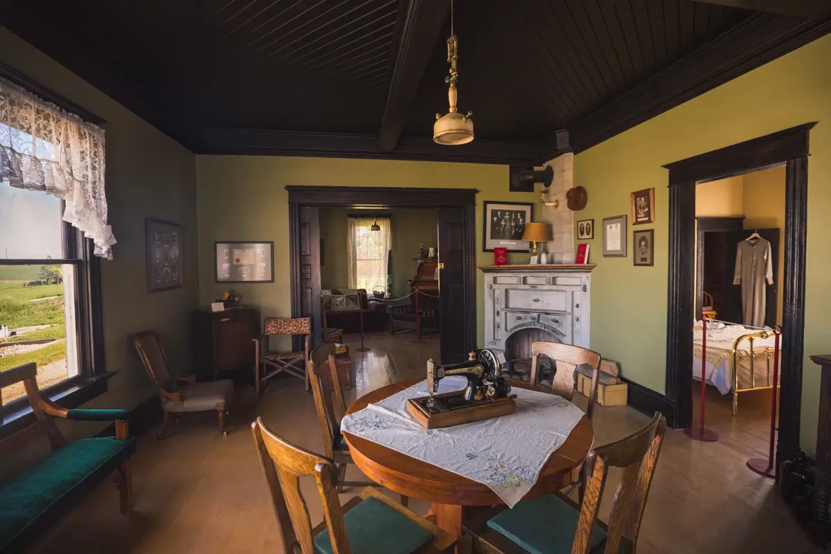 A vintage Lethbridge room with wooden furniture, a round table with a sewing machine, green sofa, and framed pictures on pale green walls. An adjacent bedroom with a bed and dress on a hanger is visible through an open doorway.