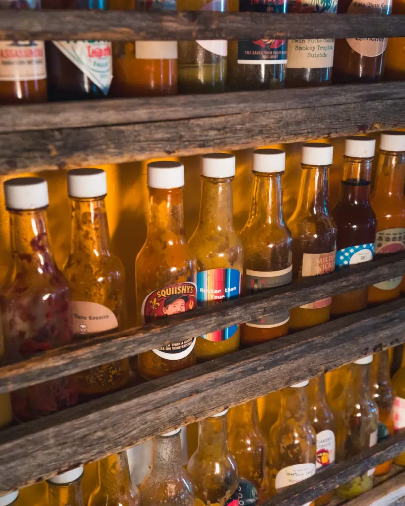 Glass bottles of various hot sauces with white caps are arranged in three rows on wooden shelves against a yellow wall, adding spicy flair to this Lethbridge display. The bottles feature different colored labels and varying amounts of sauce inside.