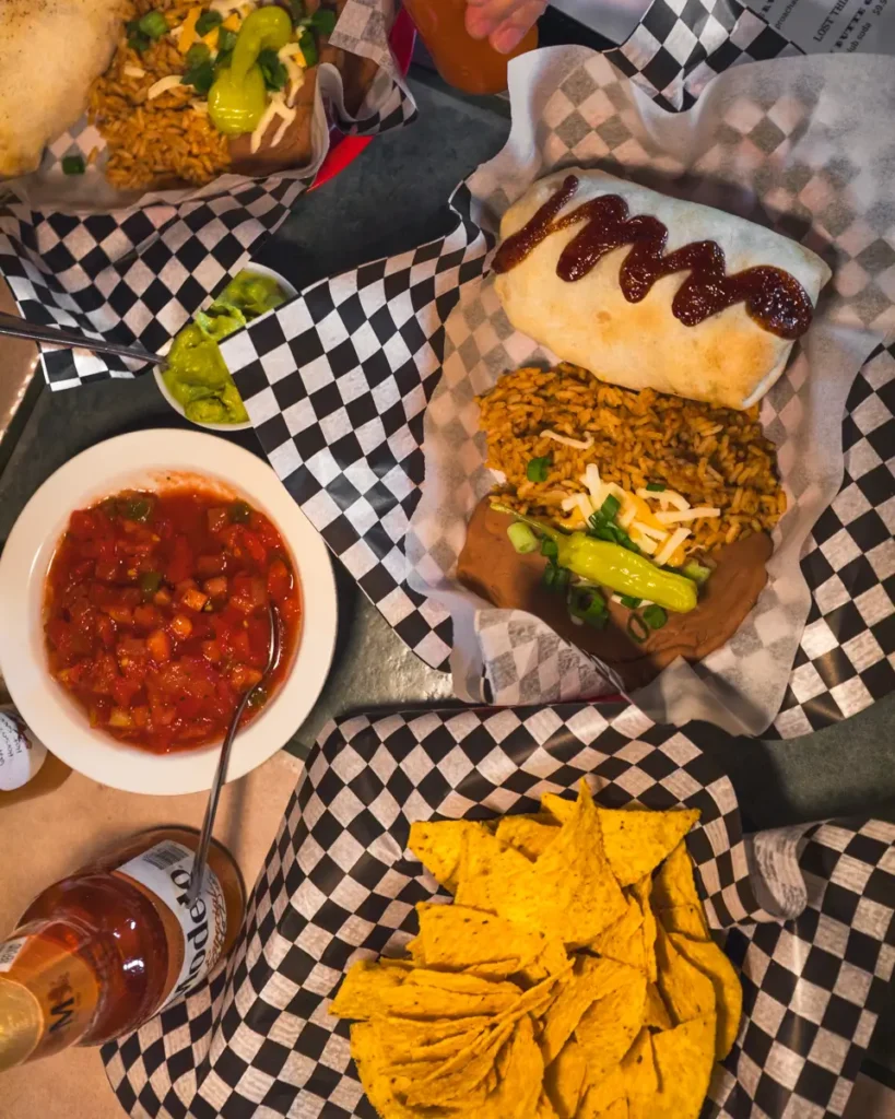 A table in Lethbridge features plates of Mexican food, including a saucy burrito, rice, sliced jalapeños, a bowl of salsa with a spoon, yellow corn tortilla chips, and bottles of hot sauce.