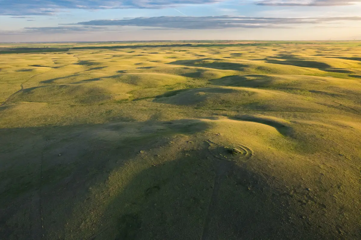 Aerial view of gently rolling grassy hills near Lethbridge under a partly cloudy sky, with sunlight casting long shadows across the landscape.