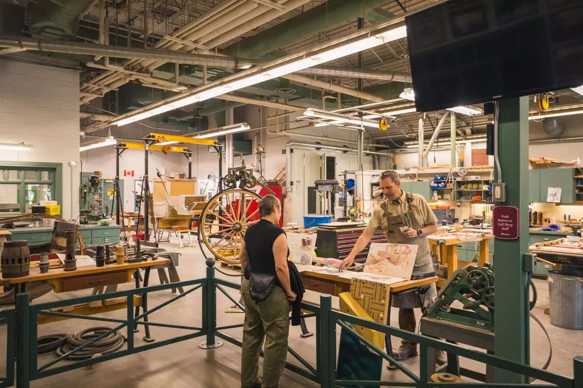 Two people stand in a Lethbridge workshop filled with tools, worktables, and a large wooden wheel. One presents drawings on a table while the other listens. The space features exposed pipes and industrial lighting.