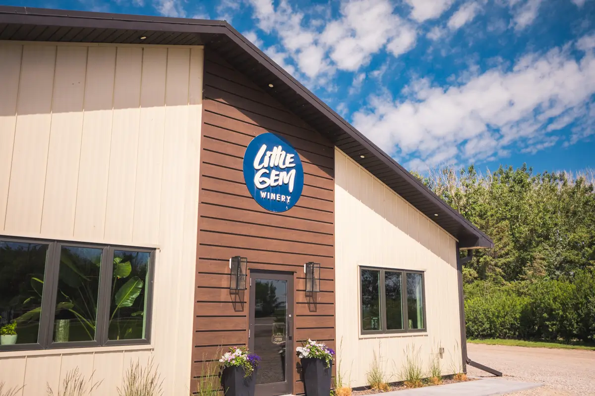 A building with cream and brown exterior siding, large windows, and a central door bears a blue circular sign reading Little Gem Winery. Set in Lethbridge, the winery stands under a clear sky with scattered clouds and trees in the background.