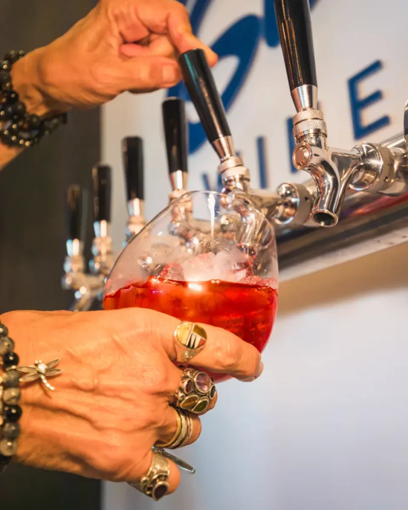 A person wearing multiple rings and bracelets fills a stemless wine glass with a red drink containing ice from a row of metal beverage taps in Lethbridge.