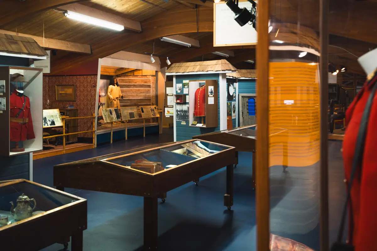 A Lethbridge museum exhibit room features display cases with historical artifacts, mannequins dressed in period clothing, informational panels, and wooden beams across the ceiling.