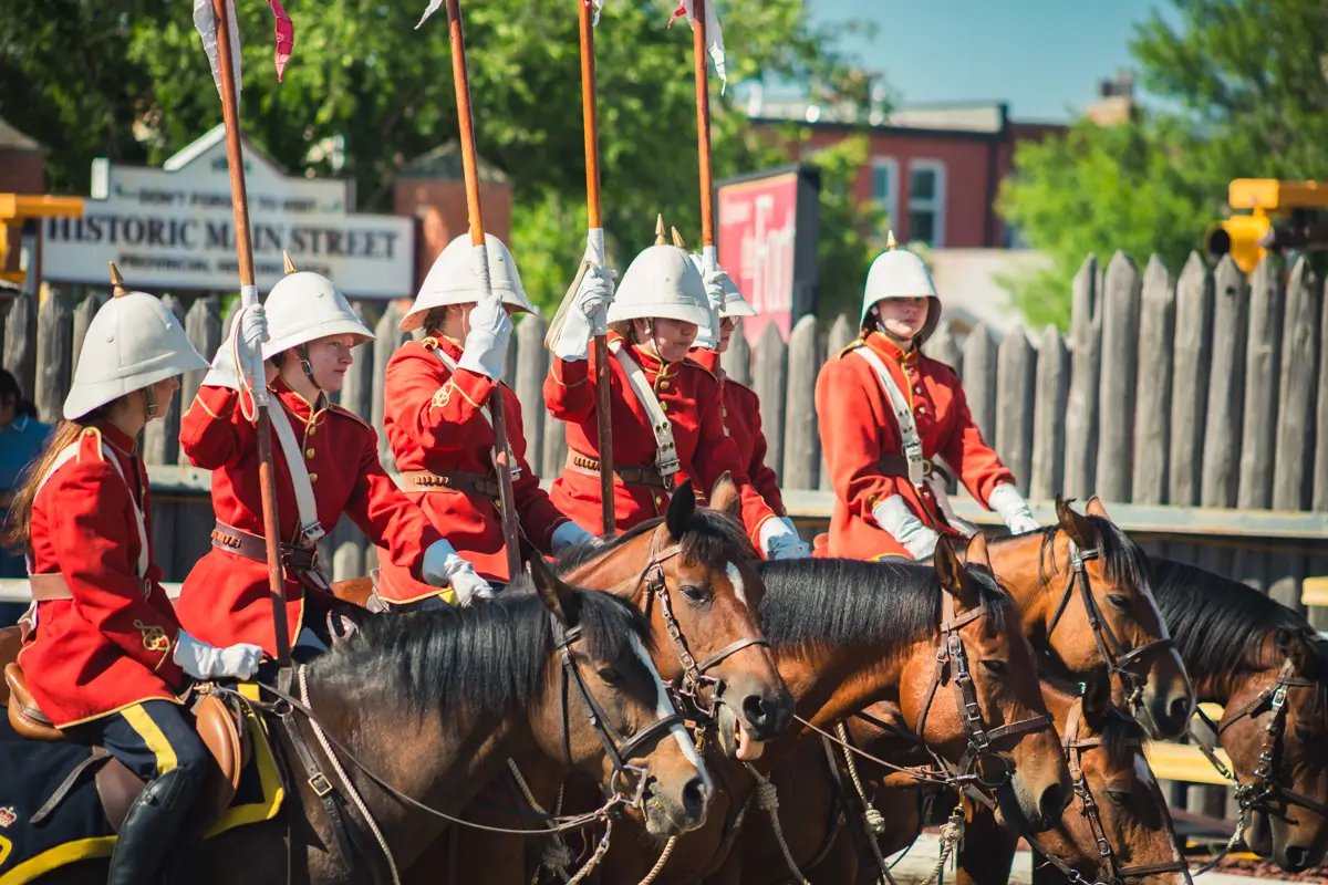 Five people in bright red uniforms and white helmets sit on horses, holding lances upright. Wooden fencing and signs reading HISTORIC MAIN STREET place the scene in the heart of Lethbridge.