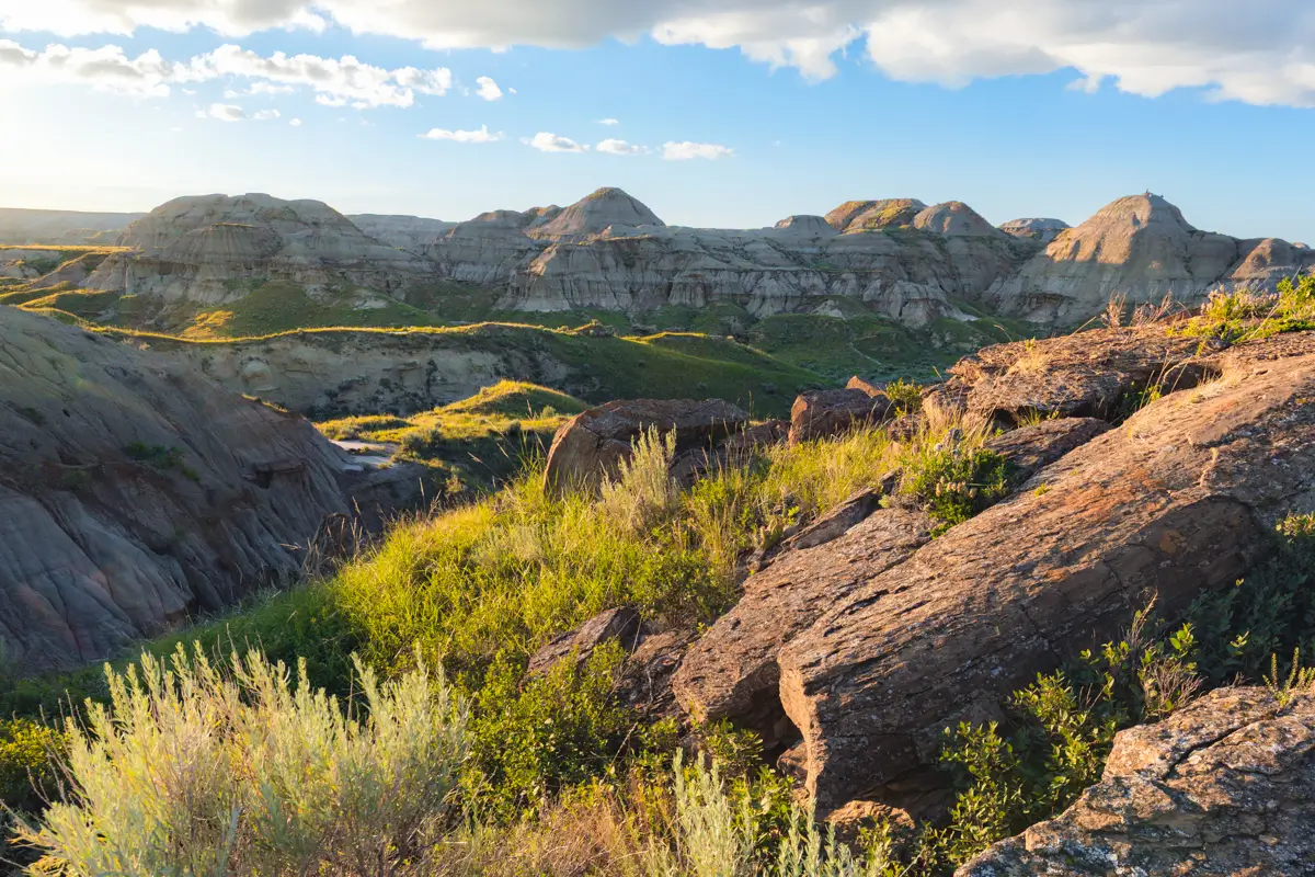 Rocky badlands landscape near Lethbridge, with layered hills in the distance, sparse green vegetation, and sunlight casting shadows across the terrain under a partly cloudy blue sky.