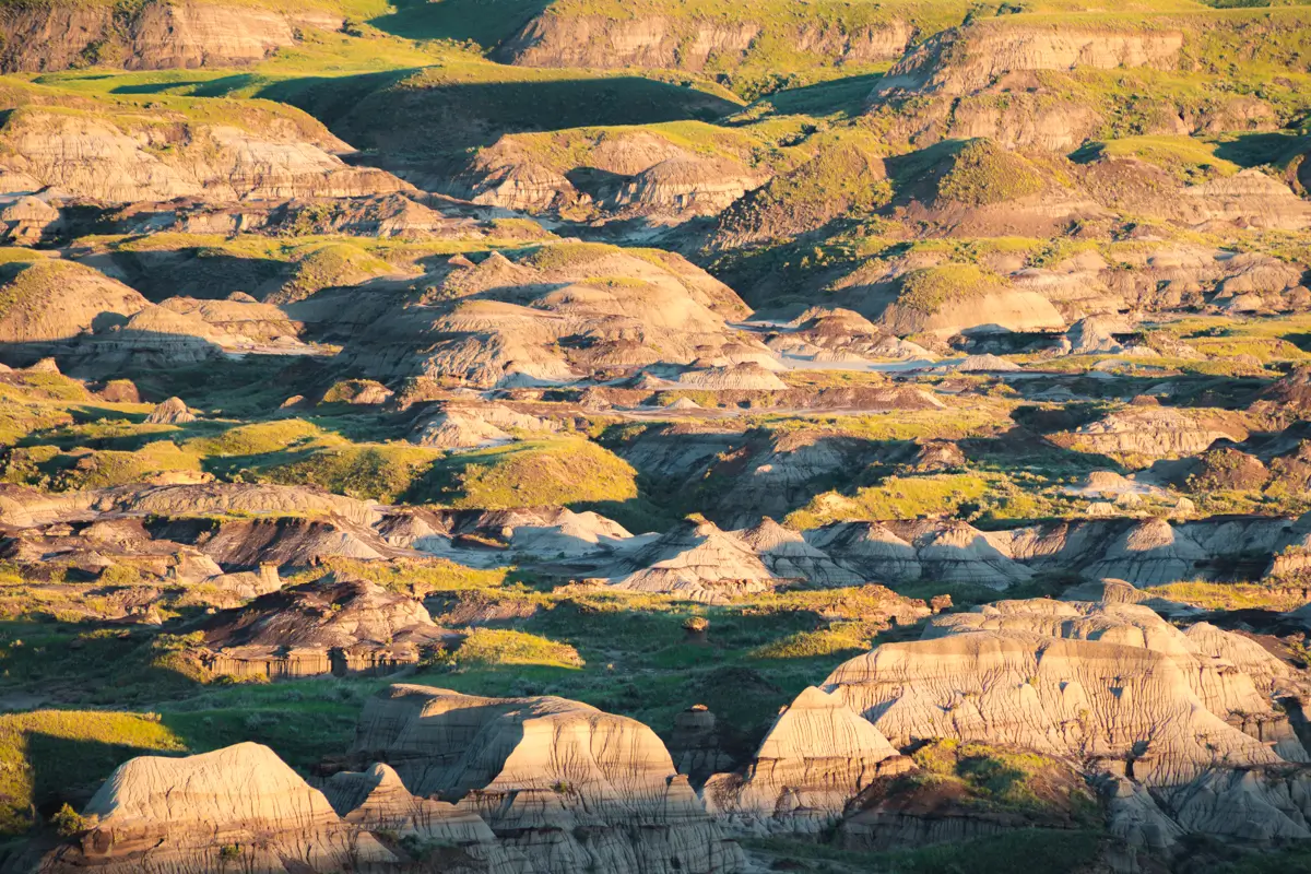 Eroded hills and rock formations with layers of sediment, bathed in sunlight, evoke the unique landscape around Lethbridge. Sparse green vegetation is scattered across the terrain, creating contrast with the rocky hills. Shadows highlight the contours of the land.