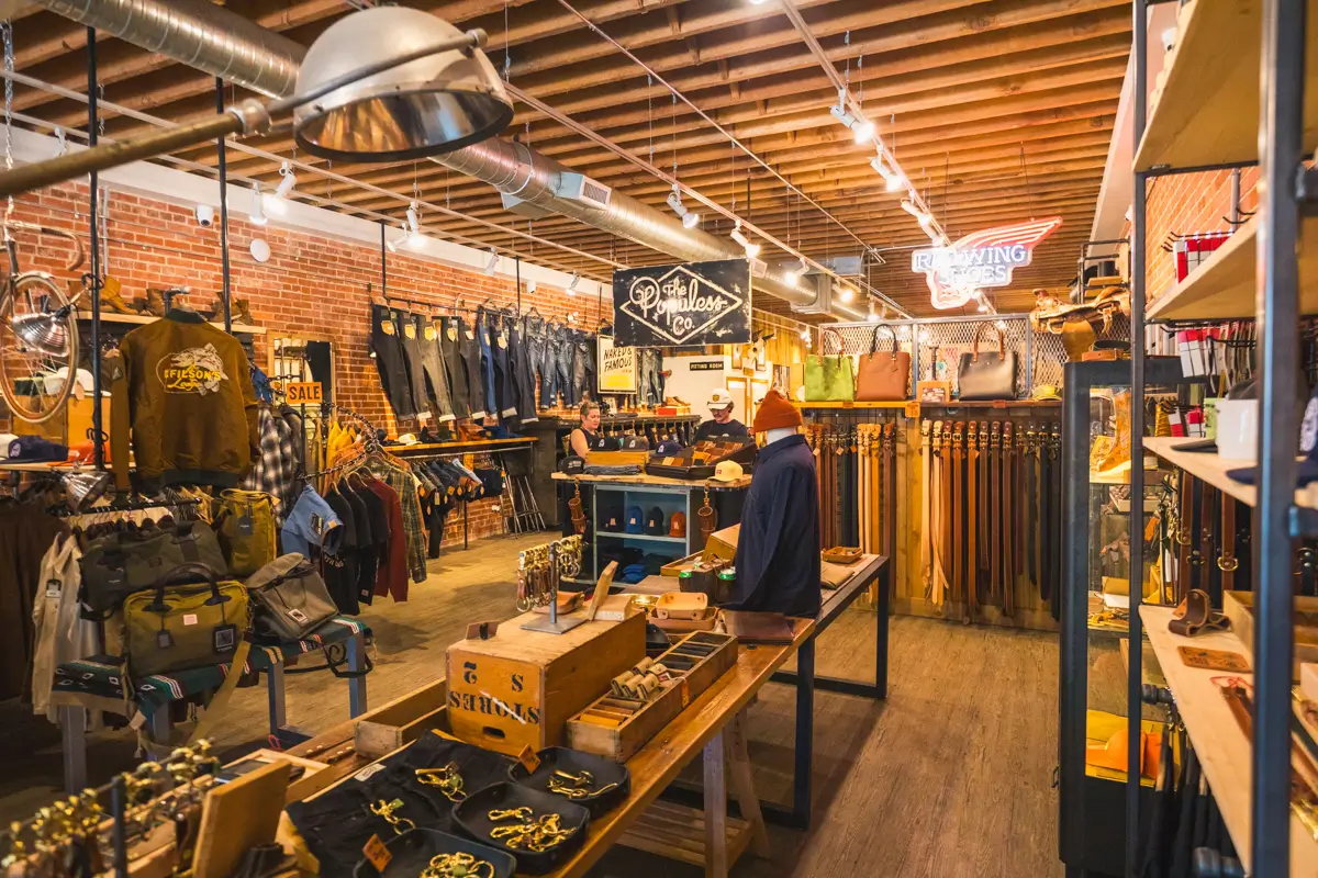 A retail store in Lethbridge with wooden floors and exposed ceiling beams, displaying clothing, belts, bags, and accessories on racks and tables. A person in a beanie stands near the center. The store has a rustic, industrial style.