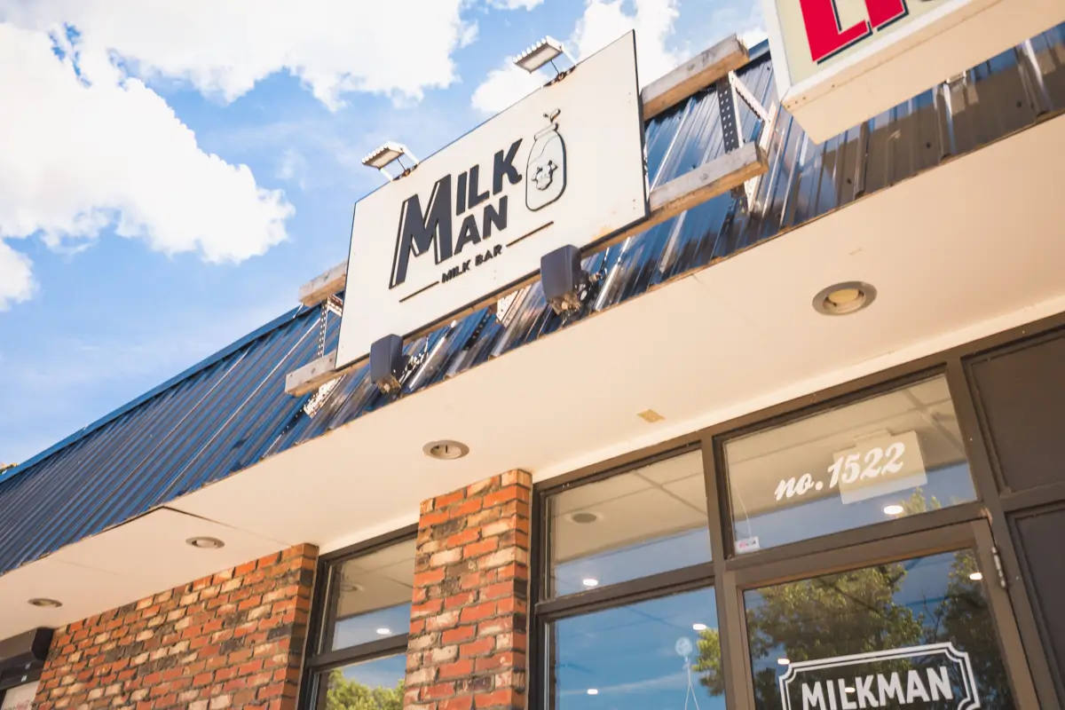 A storefront with a brick facade and large windows displays a sign reading Milk Man Milk Bar with a milk bottle graphic in Lethbridge. The address number 1522 is visible above the entrance. The sky is partly cloudy.