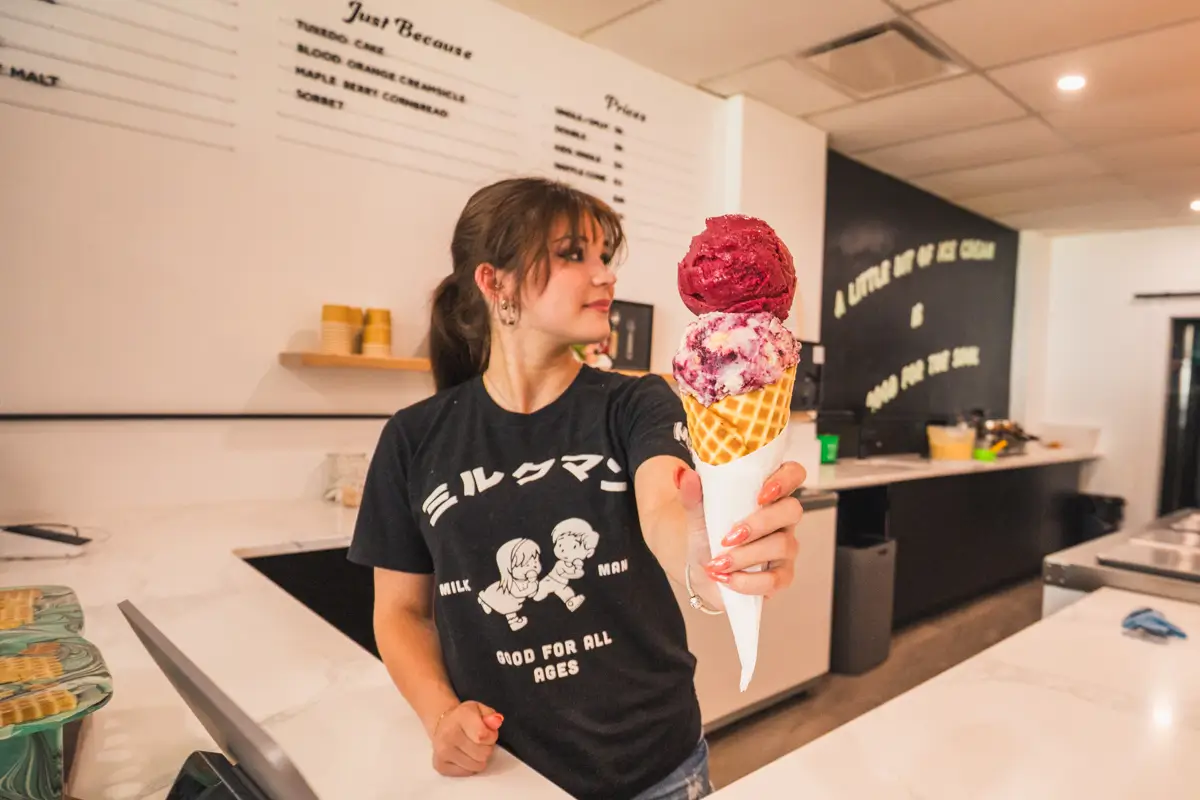 A woman behind an ice cream counter in Lethbridge holds out a waffle cone with three large scoops of ice cream. She wears a black t-shirt and stands in a modern, brightly lit shop.
