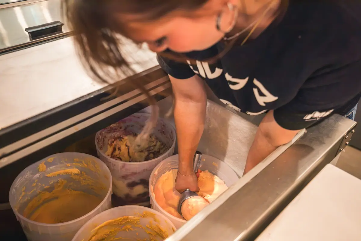 A person in Lethbridge scoops orange and white ice cream from a large tub in a freezer, surrounded by other tubs of different ice cream flavors.