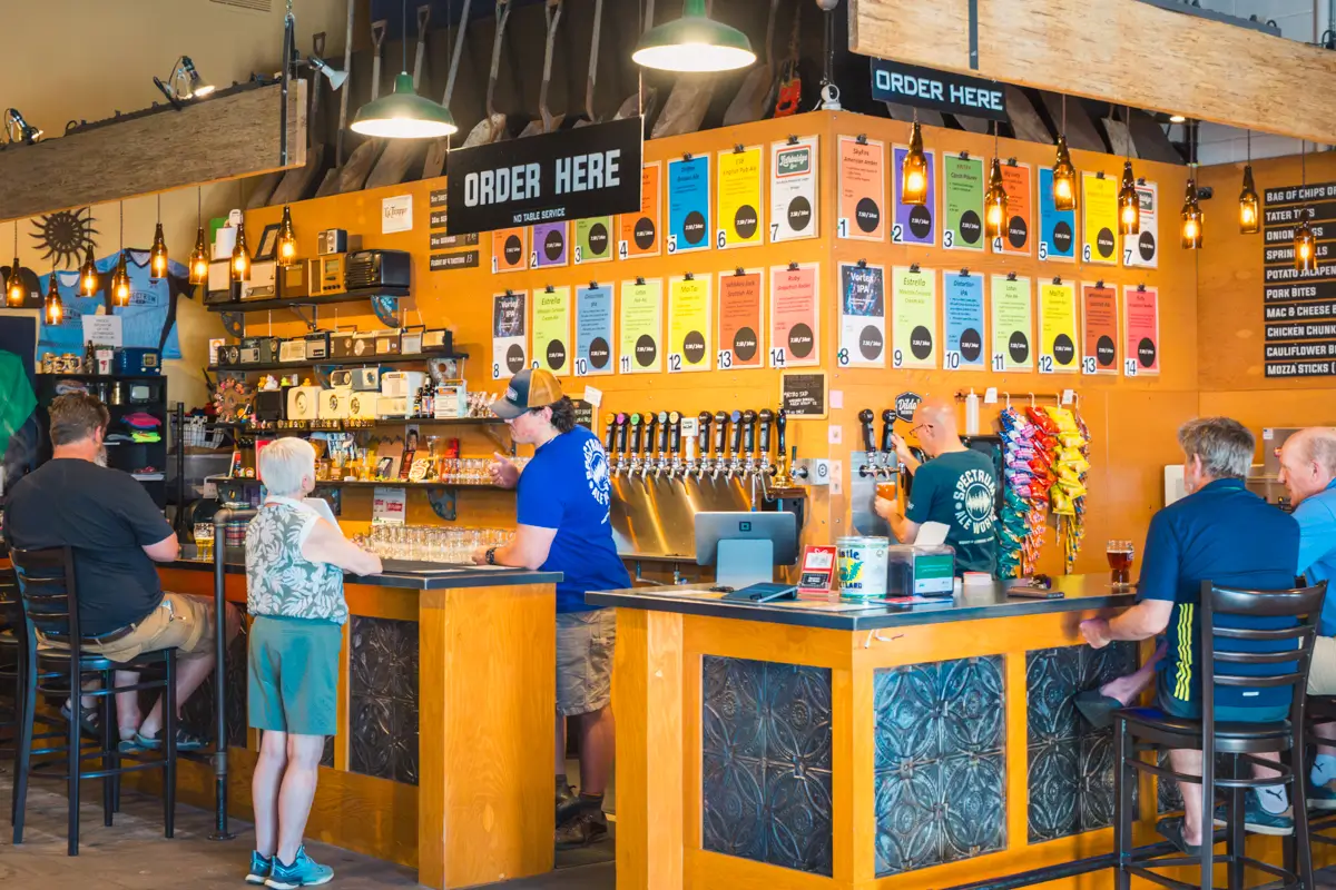 A brewery taproom in Lethbridge with people at the bar. Colorful beer menus hang above many beer taps. Pendant lights and decorations are visible. Staff serve drinks as patrons sit or stand at the counter.