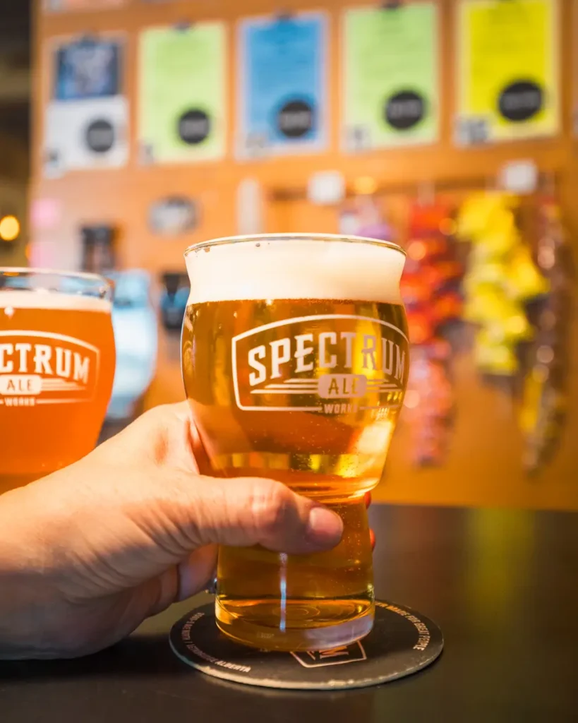 A hand holds a pint glass of golden beer labeled SPECTRUM ALE on a bar counter in Lethbridge, with another beer beside it; colorful menus and decorations are blurred in the background.