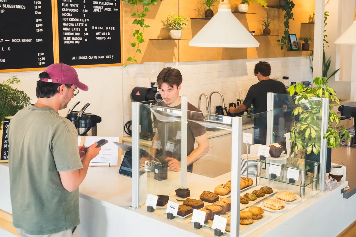 A man stands at the counter of a modern Lethbridge bakery café, looking at his phone. Behind the glass, various pastries are displayed. Two employees work behind the counter with plants and a menu board visible on the wall.