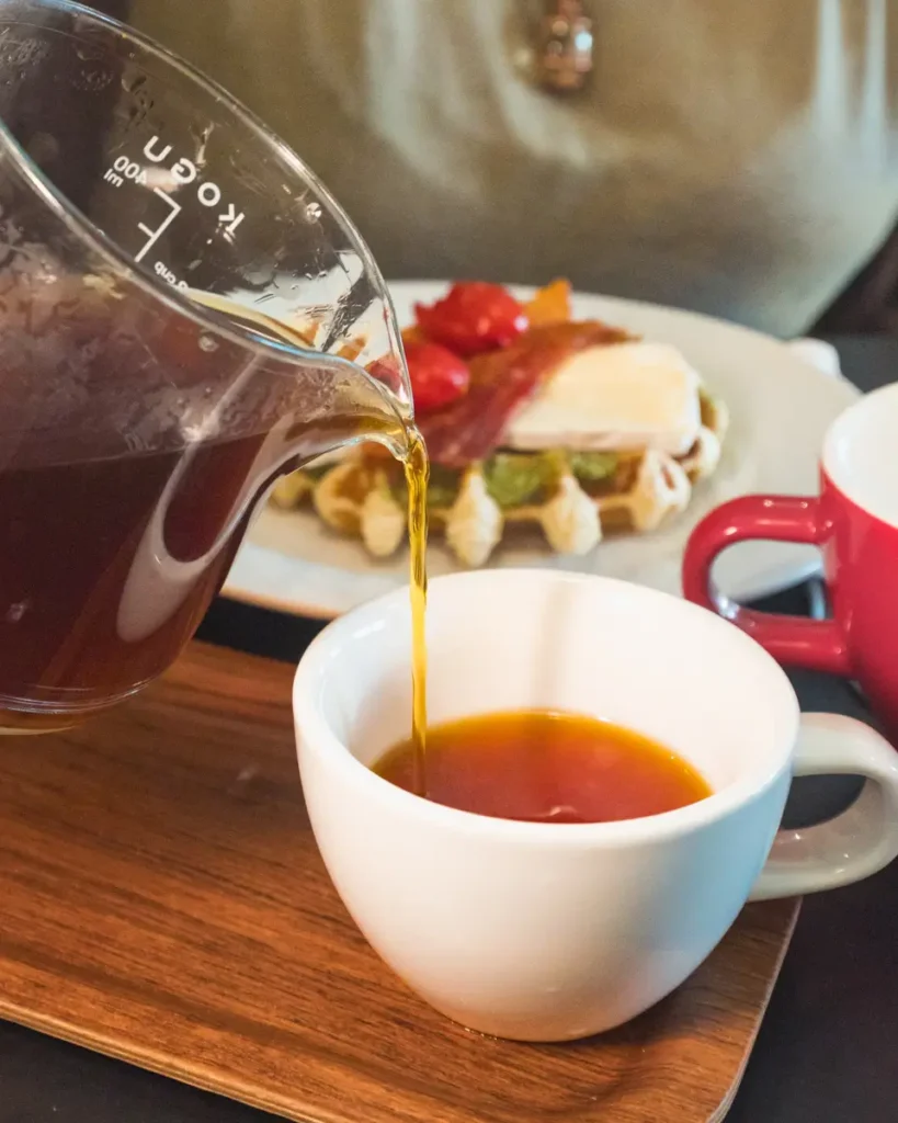 Coffee is being poured from a glass carafe into a white cup on a wooden tray, creating an inviting scene reminiscent of a cozy Lethbridge café. In the background, a plate of food with waffles, tomatoes, bacon, and avocado is partially visible. A red mug sits next to the white cup.