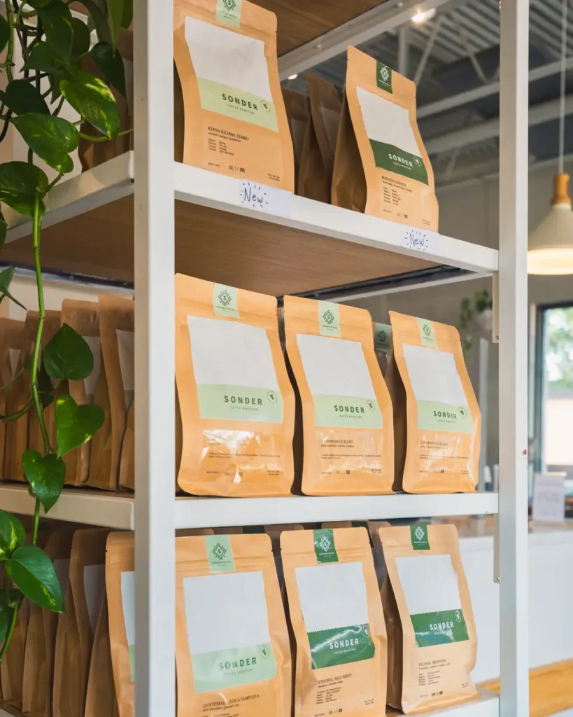 Packets of coffee labeled Sonder are neatly arranged on white shelves in a bright, modern store in Lethbridge. Green plants decorate the shelving, and sunlight comes through large windows in the background.
