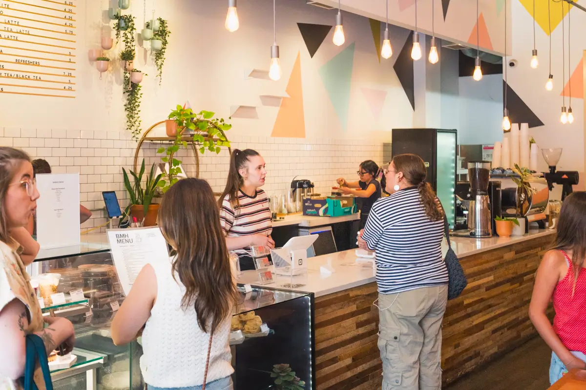 People order and interact at a modern Lethbridge coffee shop counter with hanging lights, geometric wall art, and a wooden counter. Staff members prepare drinks and assist customers while pastries are displayed in a glass case.
