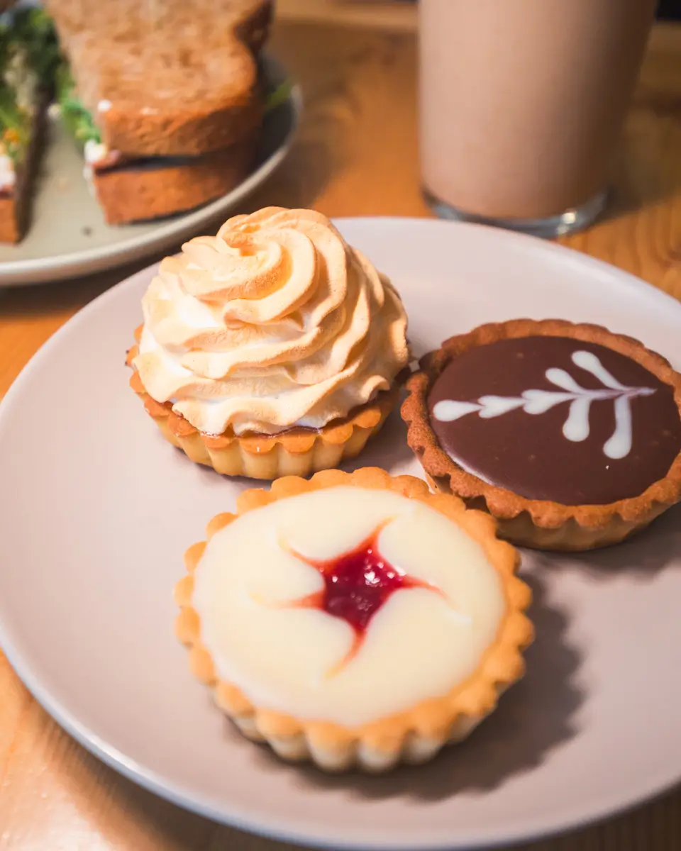 A plate with three tarts—one with whipped topping, one with chocolate and white icing, and one with a red jam center—showcases a delightful Lethbridge treat. In the background, a sandwich and a glass of light-colored drink complete this cozy scene.